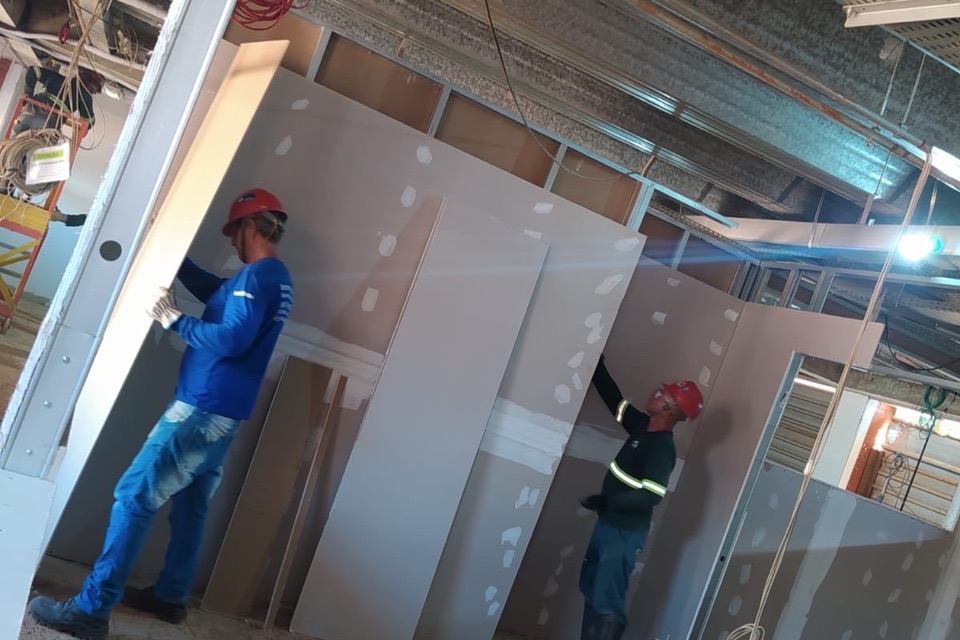 Equipe técnica instalando painéis de drywall em obra, com estrutura de forro e sinalização de segurança ao fundo.