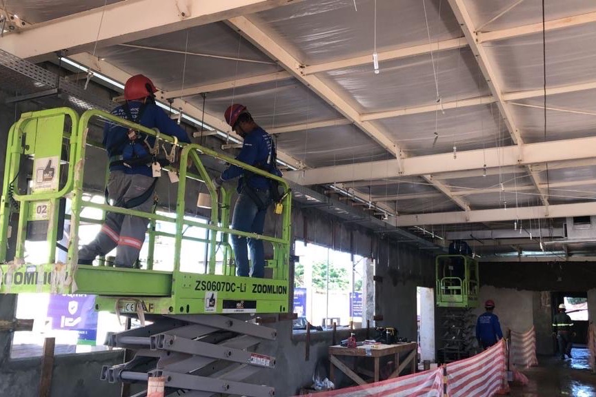 Equipe técnica em plataforma elevatória, instalando infraestrutura elétrica suspensa em teto industrial, com uso de EPI.