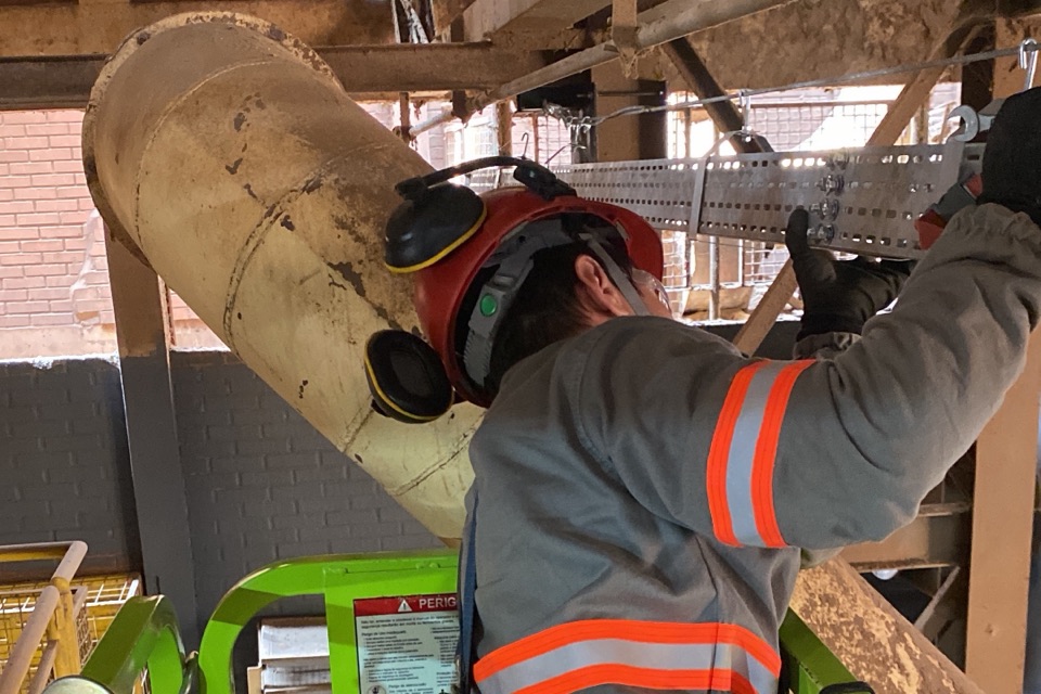 Técnico com capacete, protetores auriculares e jaqueta com faixas refletivas fixando suporte de calha metálica em duto industrial, trabalhando em plataforma elevatória.