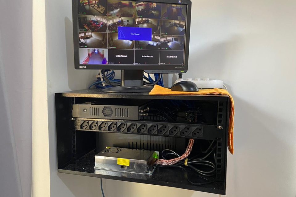 Monitor de CFTV com mosaico de câmeras sobre rack de parede com gravador.