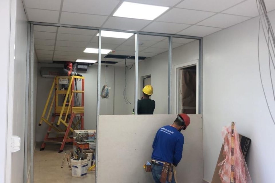 Equipe da INOVE, com capacetes e camisas da empresa, montando divisórias de drywall em escritório em obra.