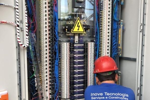 Técnico da INOVE, de capacete vermelho e camisa da empresa, trabalhando diante de um grande painel elétrico com fiação organizada.