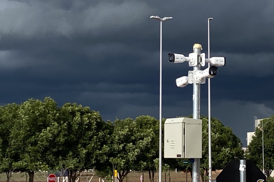 Conjunto de câmeras de CFTV instaladas em poste metálico, com caixa de conexão, sob céu de tempestade.